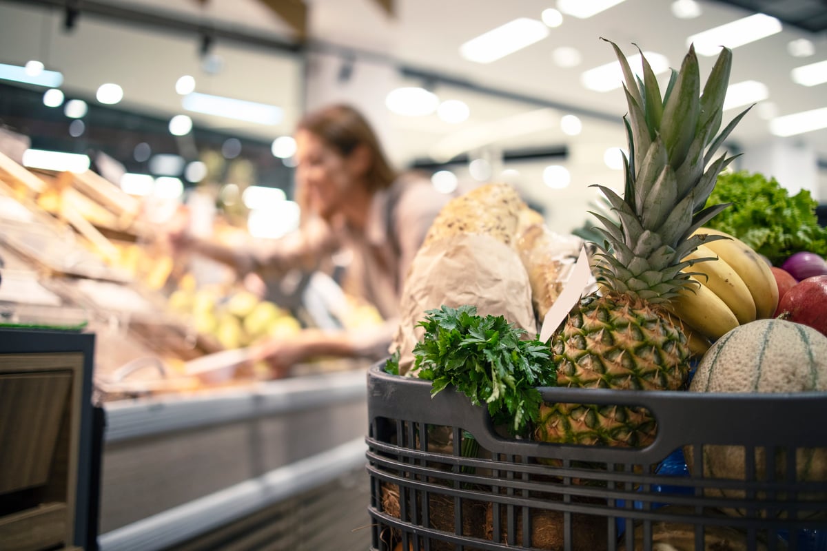 closeup-shopping-cart-supermarket-full-food-fruit-vegetables-while-background-woman-taking-product-off-shelves