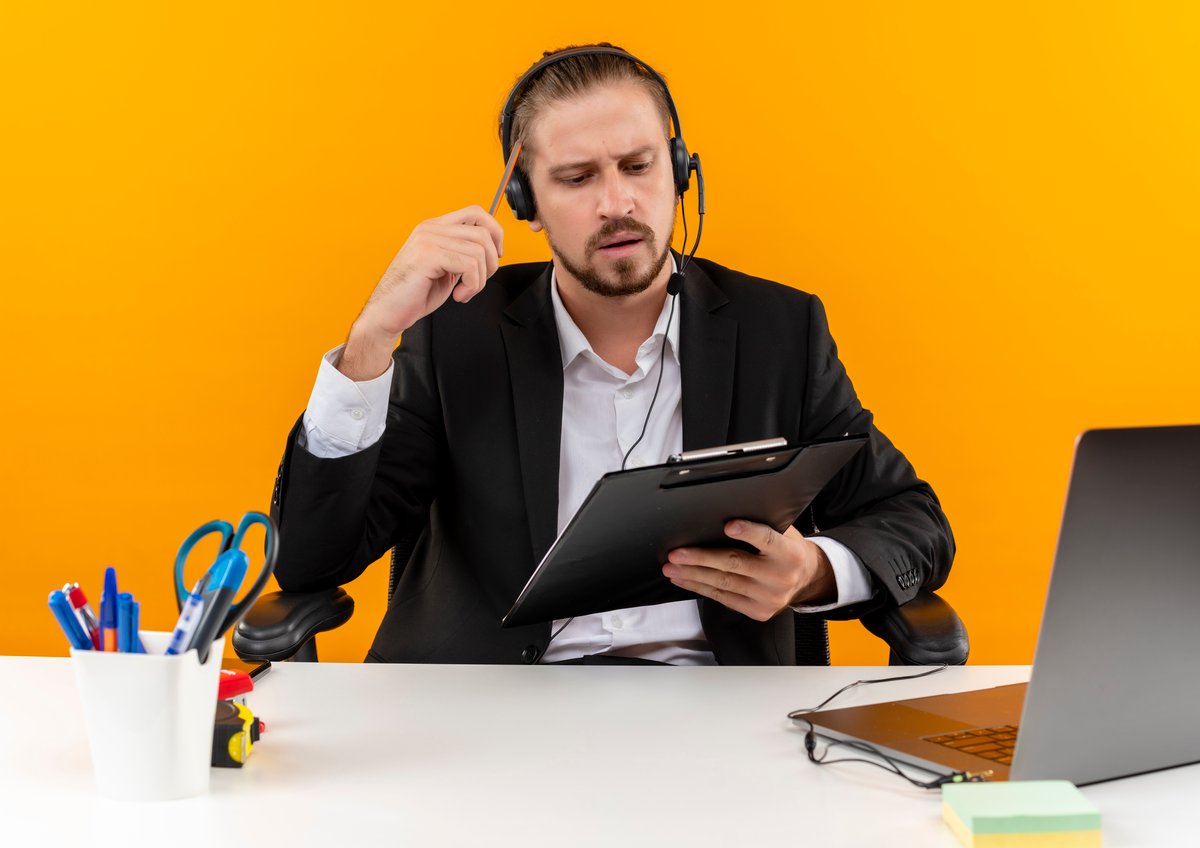 handsome-business-man-suit-headphones-with-microphone-holding-clipboard-looking-it-with-serious-face-sitting-table-offise-orange-background